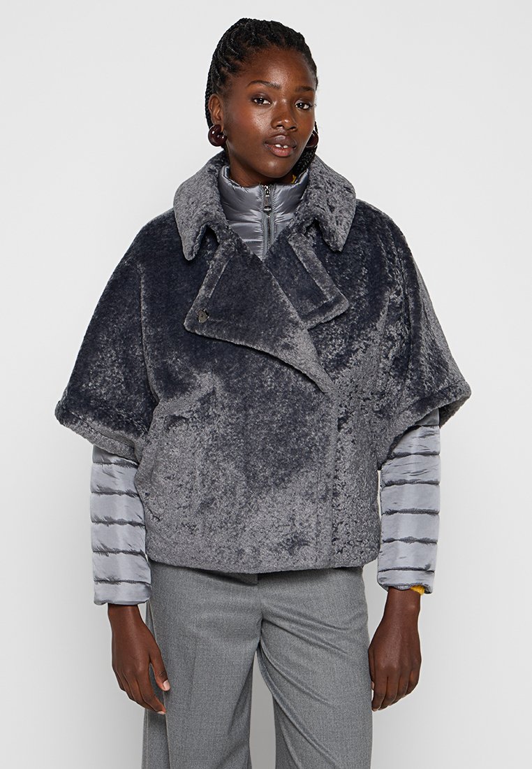 Schwarze Frau, die einen geschichteten grauen texturierten Kurzpelzmantel über einer grauen wattierten Jacke und grauen Hosen trägt, vor einem schlichten Hintergrund steht.