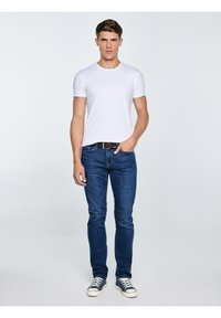 Fiatal férfi fehér pólóban, kék farmerben, fekete övvel és kék tornacipőben, egyszínű világos háttér előtt áll.