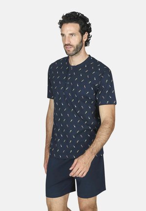 Uomo con capelli ricci che indossa una camicia blu navy a maniche corte con piccoli motivi multicolori e pantaloncini blu navy, in piedi davanti a uno sfondo bianco.