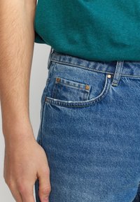 Blauwe spijkerbroek van denim met een vervaagde textuur, voorzien van twee zijzakken, gouden stiksels en bronzen klinknagels, gedragen met een groene T-shirt.