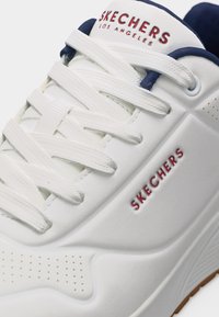 Λευκό αθλητικό παπούτσι Skechers με λευκά κορδόνια, κόκκινο κείμενο "SKECHERS LOS ANGELES" στη γλώσσα και στο πλάι, και επένδυση σε σκούρο μπλε.