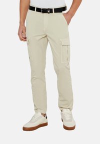Pantalones cargo beige con un corte recto, cinturón de tela, dos bolsillos laterales y dos bolsillos cargo. Combinados con zapatillas blancas y suelas de goma claras.