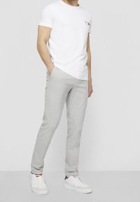 White short-sleeve t-shirt with a small logo on the chest, paired with light grey, slim-fit trousers and white sneakers. Simple design and colors.