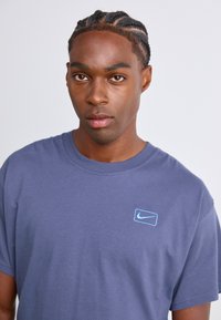 Ung mand med fletninger iført en blå Nike t-shirt, der kigger direkte frem mod en ensfarvet lys baggrund.