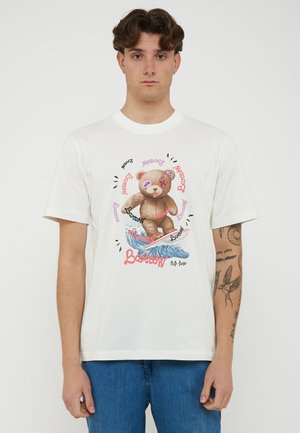 Giovane uomo che indossa una t-shirt bianca con un orsetto surfista su un'onda e la scritta "Barstow" in più colori.