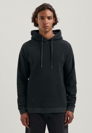 Jong persoon met middellang krullend haar, gekleed in een donkergrijze hoodie en donkere broek, staand tegen een effen achtergrond.