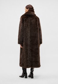 Dlhý hnedý kožuch s vysokým golierom, majúci hustú textúru a rovný strih. Nosený s čiernymi čižmami po kolená.