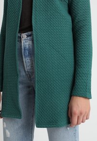 Tyrkysový, strukturovaný cardigan s tkaným vzorem, otevřený vpředu a s dlouhými rukávy. Nosený přes černý top a světle modré, poničené džíny.