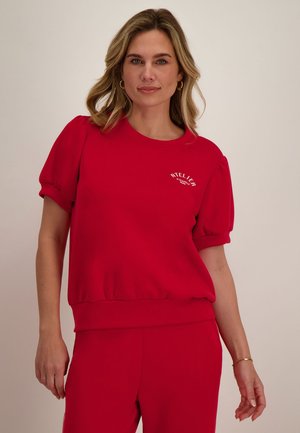 Vrouw met schouderlang blond haar draagt een rood kort mouwen top met pofmouwen en bijpassende rode broek, staand tegen een effen achtergrond.