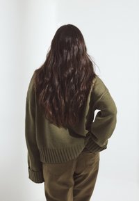 Olive green knit sweater with ribbed cuffs, relaxed fit, paired with matching green pants. Long wavy hair visible at the back.