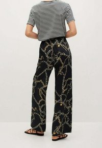 Persoon in zwarte wijde broek met gouden kettingprint, gestreept zwart-wit t-shirt met korte mouwen en zwarte sandalen met banden, staand.