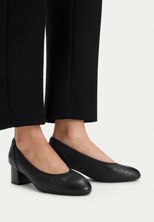 Schwarze, flache Lederschuhe mit dezentem, perforiertem Blumenmuster, getragen zu schwarzen Hosen vor weißem Hintergrund.