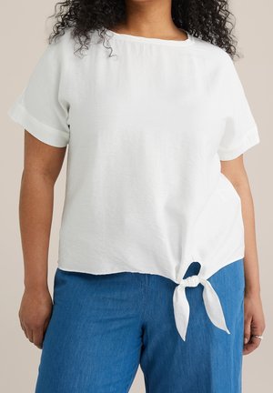 Kvinde iført en hvid kortærmet bluse med en knude foran, kombineret med blå bukser, mod en neutral baggrund.