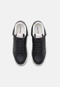 Para czarnych niskich sneka z białymi podeszwami, czarnymi sznurówkami, logo Emporio Armani na języku i boku, widziane z góry na białym tle.