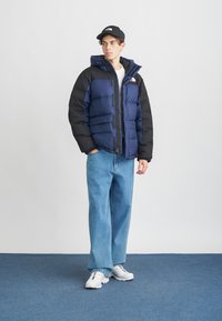 Noor noormees, kellel on seljas sinine ja must sulejakk, heledad sinised teksad, valged tossud ja must müts, seisab ühe käe taskus.