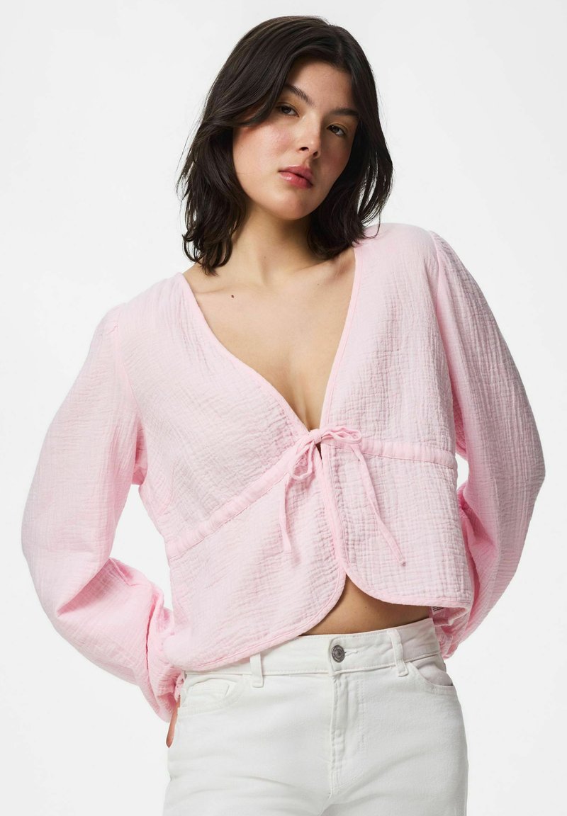 Femme aux cheveux foncés mi-longs portant un chemisier rose clair texturé noué devant et un pantalon blanc taille haute sur fond uni.