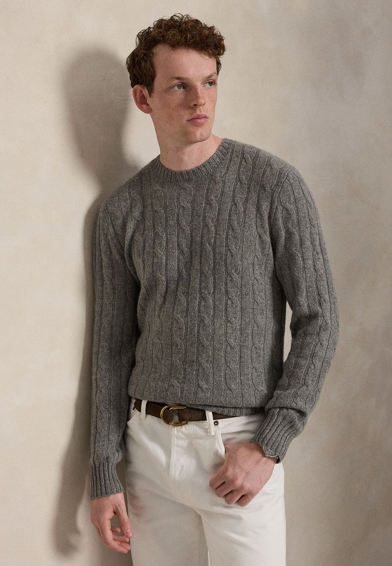 Junger Mann mit lockigem braunem Haar, der einen grauen Zopfpullover, weiße Hose und einen braunen Gürtel trägt, steht vor einer beigen Wand.