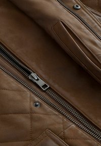 Veste en cuir marron avec un design matelassé, fermeture éclair en argent, boutons-pression assortis et une texture lisse. Comprend une poche latérale.