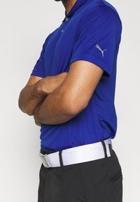 Polo bleu à manches courtes avec col, arborant un logo discret sur la manche gauche. Associé à une ceinture blanche et un pantalon noir.