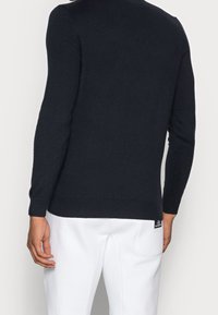 Maglione in maglia blu navy con maniche lunghe, polsini e orlo a coste, e una texture liscia. Indossato con pantaloni bianchi, vista posteriore.