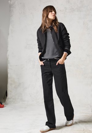 Veste zippée noire par-dessus un pull à motifs noir et blanc, associée à un jean large noir et des baskets blanches. Texture lisse, coupe décontractée.