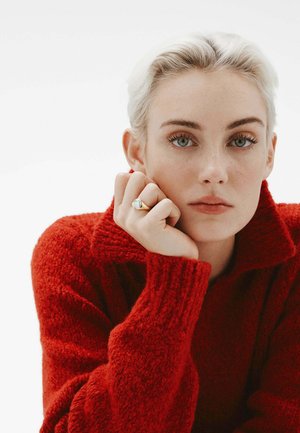 Jersey texturizado rojo con cuello alto. El sujeto tiene el cabello corto y claro, y lleva un anillo dorado con una piedra clara en su mano izquierda.