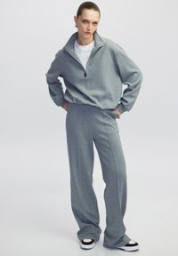 Grijze zip-up sweatshirt gecombineerd met grijze wijde broek. De stof lijkt zacht, met een relaxte pasvorm en casual ontwerp. Zwart-witte sneakers.