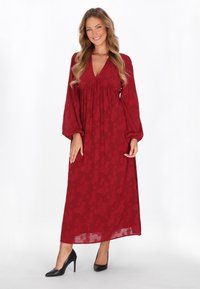 Vestido largo rojo con escote en V profundo, mangas fluidas y patrón floral texturizado. Llevado con tacones negros sobre un fondo neutro.