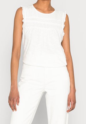 Mouwloze witte blouse met geruite randen en getextureerde horizontale strepen, gecombineerd met witte broek met hoge taille op een staand persoon.