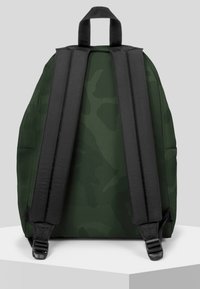 Mochila de camuflaje verde con correas negras y asa superior. Presenta un tejido suave, forma rectangular y hebillas laterales ajustables.