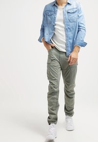 Ljusblå denimskjorta, vit t-shirt med rund hals, gröna cargobyxor med fickor och elastiska manschetter, samt vita sneakers. Avslappnad outfit för män.