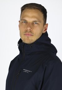 Marineblaue wasserdichte Jacke mit Kapuze, ausgestattet mit einem Frontreißverschluss, verstellbaren Schnüren und einem Logo auf der oberen linken Seite. Glatte Textur.