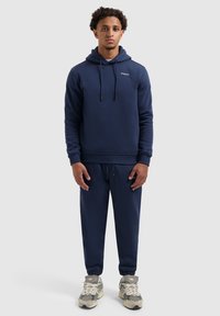 Jongeman staat met een neutrale gezichtsausdruk, gekleed in een marineblauwe hoodie en bijpassende joggingbroek, met grijze sneakers op een effen lichte achtergrond.
