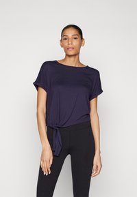 Mørkeblå t-skjorte med rund hals, korte ermer og knutetaille i livet, sammen med svarte leggings. Glatt stofftekstur.