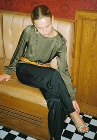 Grön sidenblus med draperade ärmar, svarta satinbyxor och silverfärgade sandaler. Sitter på en beige bänk mot en röd vägg.