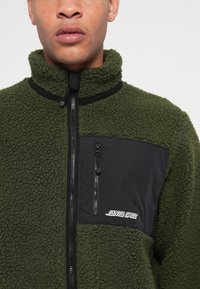 Fleecejakke i mørkegrønn med lomme i svart nylon på brystet, glidelåslukking og logo-detalj; teksturert ytterside og høy halsdesign.
