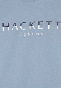 Heled sinise puuvillase T-särgi, millel on boldne ja kontrastne must-valge tüpograafia tekstidega "HACKETT" ja "LONDON". Sujuv tekstuur.