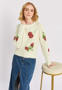Biały puszysty sweter z haftowanymi wzorami wiśni w czerwonym brokacie i zielonymi akcentami, zestawiony z niebieską dżinsową spódnicą z rozcięciem z przodu.
