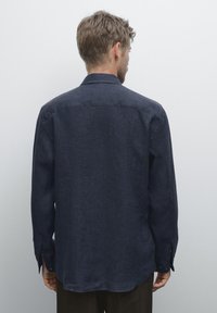 Man with short brown hair wearing a dark blue long-sleeve shirt, viewed from behind against a plain light background.