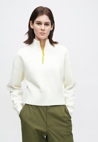 Mladá osoba s tmavými vlasy do ramen, oblečená v bílém svetr s půlzipem a žlutým zipem, doplněná olivovými kalhotami, se rukama v kapsách.