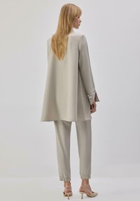 Ensemble de costume gris clair comprenant un blazer long et ample ainsi que des pantalons fuselés avec des poignets élastiques, fabriqué dans un tissu lisse, associé à des talons minimalistes.