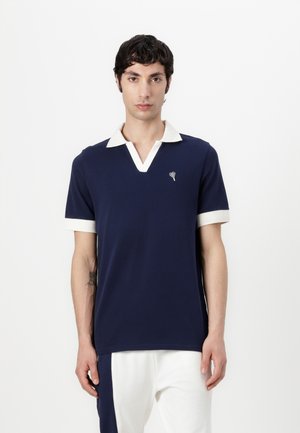 Polo de color navy con cuello y puños de manga blancos. Presenta un escote en V y un pequeño diseño de corazón bordado en el pecho. Textura de tela suave.