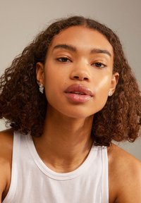 Pendientes de plata con diseño de hoja y acentos de piedras preciosas, llevados por un modelo con cabello rizado, que viste una camiseta sin mangas blanca de canalé.