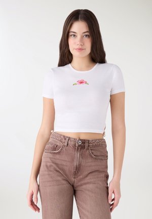 Giovane donna che indossa una maglietta bianca corta con ricamo di fiori rosa e jeans marroni a vita alta, in piedi davanti a uno sfondo semplice.