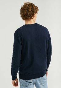 Pull en maille bleu marine à manches longues, encolure ronde, poignets et ourlet côtelés, porté avec un jean bleu clair, vu de dos.