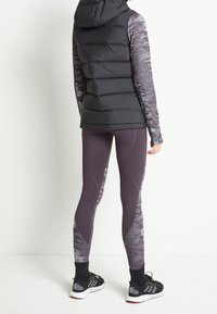 Persona con chaleco acolchado negro, camiseta de manga larga con estampado, mallas moradas con estampado y zapatillas deportivas negras, de pie contra un fondo blanco.