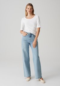 Chemise blanche à manches trois-quarts associée à un jean large bleu clair, avec passants de ceinture et détails cousus. Les chaussures sont des mules couleur crème.