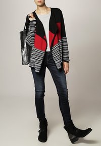 Cardigan a strisce nere e rosse, camicia grigia sotto, abbinato a jeans scuri e stivaletti neri alla caviglia. Maglia a maglia strutturata con un design a strati.