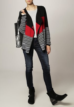 Cardigan a strisce nere e rosse, camicia grigia sotto, abbinato a jeans scuri e stivaletti neri alla caviglia. Maglia a maglia strutturata con un design a strati.