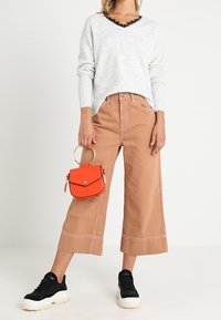 Maglione grigio con bordo in pizzo, pantaloni larghi color beige, sneakers nere e una piccola borsa arancione con manico circolare.